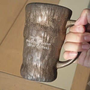 Puede incluir: Una taza cil&iacute;ndrica de cer&aacute;mica con un acabado met&aacute;lico de color bronce oscuro texturizado que recuerda a la corteza de un &aacute;rbol. La taza presenta un texto impreso en blanco: Michael - Morning Person (After Coffee). Tiene un asa curva a la derecha.