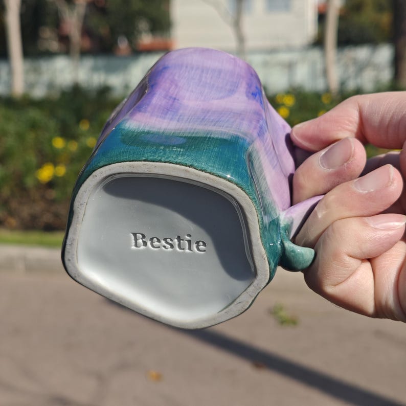 May include: A ceramic mug with a unique shape and a handle. The mug features a blend of purple, teal, and green colors. The word "Bestie" is embossed on the bottom of the mug.