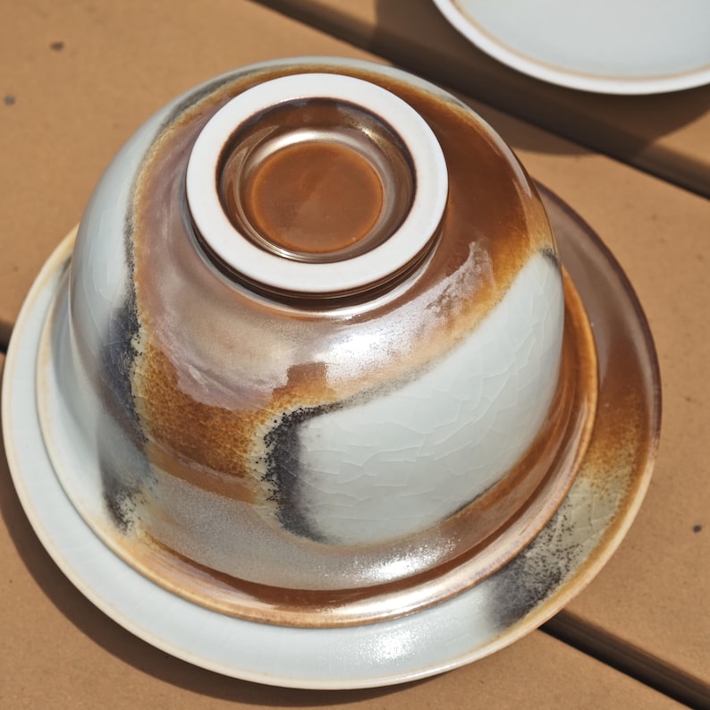 May include: A ceramic bowl and matching saucer set featuring a white crackle glaze finish with blended brown and charcoal grey accents. The bowl is inverted, displaying a smooth white circular base, while the saucer mirrors the same colour palette.