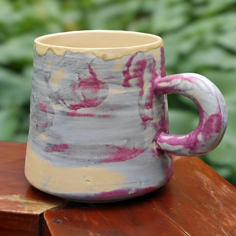 May include: Handmade ceramic mug with a unique marbled design. The mug features a cylindrical shape with a rounded handle. The color palette includes gray, purple, and yellow hues, creating a visually appealing aesthetic. Perfect for enjoying your favorite beverage.