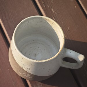 May include: A ceramic mug with a two-tone design featuring a speckled cream-coloured upper section and a solid brown lower section. The mug has a rounded handle and an embossed line drawing of a cat with the text "Hello Buddy" on the lower side.