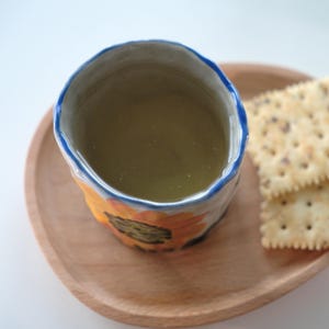 May include: A ceramic mug with a sunflower design, filled with a light green liquid, sits on a wooden tray next to two crackers. The mug has a blue rim and yellow and orange sunflower details. The tray is made of light-colored wood.