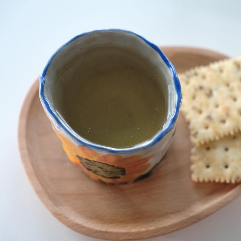May include: A ceramic mug with a sunflower design, filled with a light green liquid, sits on a wooden tray next to two crackers. The mug has a blue rim and yellow and orange sunflower details. The tray is made of light-colored wood.