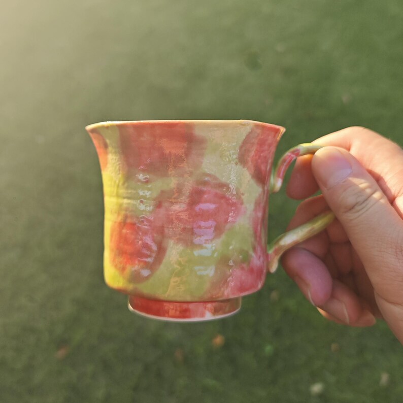 May include: A handmade ceramic mug with a unique design. The mug features a vibrant pattern of red and green splotches on a yellow background. It has a small handle and a matching red base. The mug is held in a hand against a blurred green background.