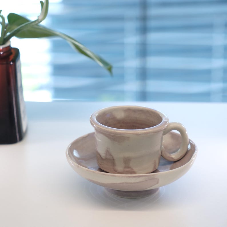 May include: A ceramic teacup and saucer set. The cup and saucer are a light beige color with brown accents. The cup has a small handle and sits on a matching saucer. The set is on a white surface.