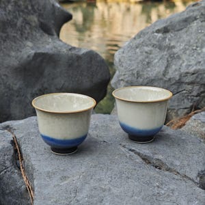 May include: Two ceramic tea cups with a cream-colored body, a blue base, and a gold rim. The cups are placed on a gray rock surface, with a blurred background of water and rocks. The cups have a minimalist design.