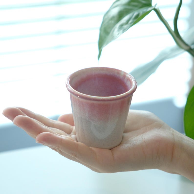 May include: A small, ceramic cup with a pink and white gradient. The cup is held in a hand, showcasing its size and design. The cup's rim is a darker pink, fading to a lighter pink and white at the base. The cup is likely for drinking.