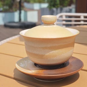 May include: A ceramic gaiwan tea set consisting of a lidded bowl and a matching saucer. The bowl features a light beige and cream horizontal striped glaze, while the saucer has a contrasting dark brown and earthy tone with a smooth finish.