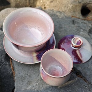 May include: A set of ceramic tea cups and saucers in shades of pink and purple. The set includes a cup, saucer, and a lidded tea bowl. The glaze has a speckled appearance, and the colors blend together. The items are arranged on a gray stone surface.