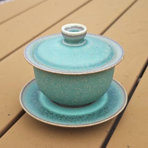 May include: A ceramic gaiwan tea set featuring a lidded bowl and a matching saucer. The set has a speckled turquoise and teal glaze with a smooth, matte finish and a dark brown rim along the edges of the lid, bowl, and saucer base.
