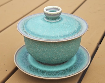 May include: A ceramic gaiwan tea set featuring a lidded bowl and a matching saucer. The set has a speckled turquoise and teal glaze with a smooth, matte finish and a dark brown rim along the edges of the lid, bowl, and saucer base.