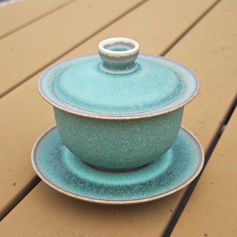 K&ouml;nnte beinhalten: Ein Gaiwan-Teeservice aus Keramik, bestehend aus einer Schale mit Deckel und einer passenden Untertasse. Das Set hat eine gesprenkelte t&uuml;rkise und blaugr&uuml;ne Glasur mit matter Oberfl&auml;che und einem dunkelbraunen Rand an Deckel, Schale und Untertasse.