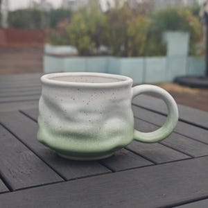 Op de afbeelding: Een handgemaakte keramische mok met een uniek, getextureerd ontwerp. De mok heeft een witte bovenkant die overgaat in een zachtgroene kleur aan de basis. Het handvat is bijpassend groen. De mok staat op een donkergrijze houten tafel.
