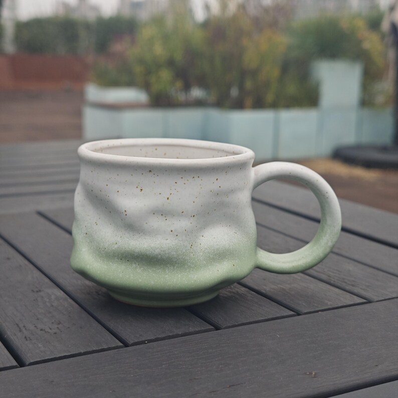 Op de afbeelding: Een handgemaakte keramische mok met een uniek, getextureerd ontwerp. De mok heeft een witte bovenkant die overgaat in een zachtgroene kleur aan de basis. Het handvat is bijpassend groen. De mok staat op een donkergrijze houten tafel.
