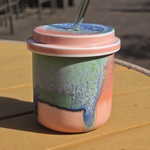 Pu&ograve; includere: Un bicchiere in ceramica con coperchio, nei toni del rosa, verde, blu e corallo. Il bicchiere &egrave; cilindrico con un coperchio dal design circolare. Una cannuccia trasparente sporge dalla parte superiore. Il bicchiere &egrave; su una superficie gialla. Altezza circa 10 cm.