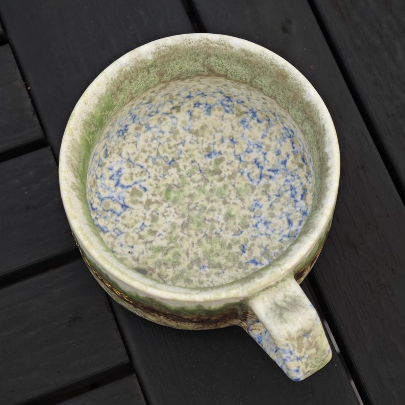May include: A ceramic bowl with a handle, featuring a speckled pattern in shades of green, blue, and cream. The bowl's rim has a green and brown border. The handle is a continuation of the bowl's design.