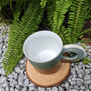K&ouml;nnte beinhalten: Eine handgefertigte Keramiktasse mit wei&szlig;em Innenraum und gr&uuml;n-blauer Au&szlig;enseite. Die Tasse steht auf einem kleinen, runden Untersetzer aus Holz. Farne und kleine graue Steine befinden sich im Hintergrund.