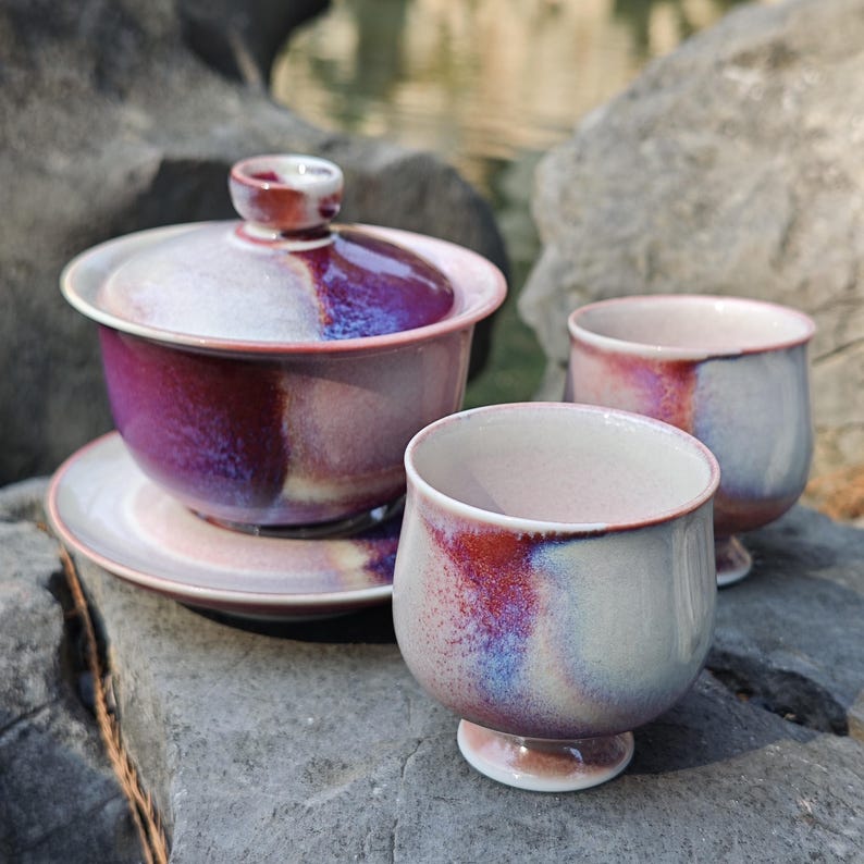 May include: A ceramic tea set with a purple and white glaze. The set includes a lidded gaiwan, a saucer, and two small teacups. The teacups have a rounded shape and a small footed base. The set is displayed on a gray stone surface.