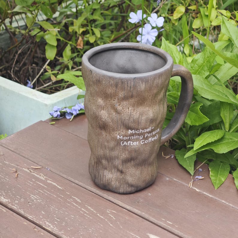 Puede incluir: Una taza de cer&aacute;mica marr&oacute;n con una superficie texturizada similar a la corteza y un interior oscuro. La taza cuenta con un asa en el lado derecho y texto impreso en blanco que dice: Michael- Morning Person (After Coffee). Forma cil&iacute;ndrica.