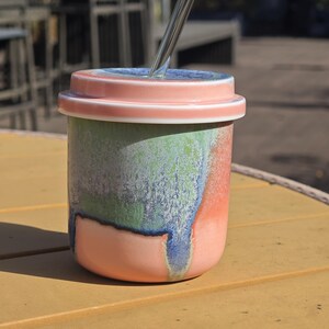 Pu&ograve; includere: Un bicchiere in ceramica con coperchio e cannuccia. Il bicchiere &egrave; cilindrico con un gradiente di colore rosa, verde e blu. Il coperchio &egrave; rosa con un bordo bianco. La cannuccia &egrave; argentata.
