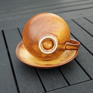 May include: A brown and yellow ceramic teacup and saucer set. The cup is round with a handle and is upside down on the saucer. The saucer and cup have a speckled design. The bottom of the cup has the text "Made in Love, Handle with Care."