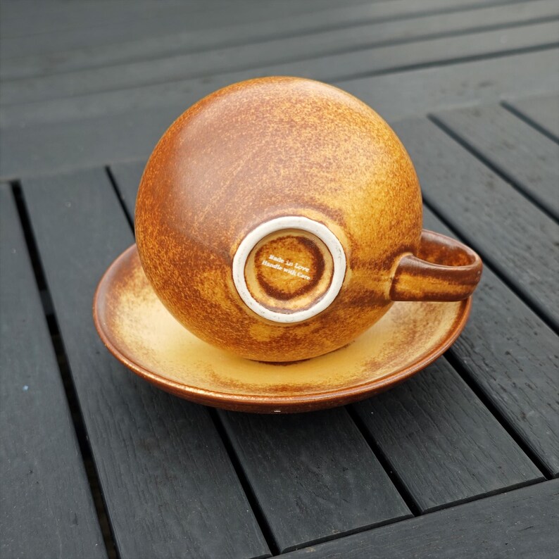 May include: A brown and yellow ceramic teacup and saucer set. The cup is round with a handle and is upside down on the saucer. The saucer and cup have a speckled design. The bottom of the cup has the text "Made in Love, Handle with Care."