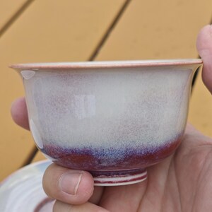 May include: A ceramic teacup with a white and purple glaze. The cup has a rounded shape with a flared rim and a small foot. The glaze transitions from white to purple near the base. The cup is held in a hand.