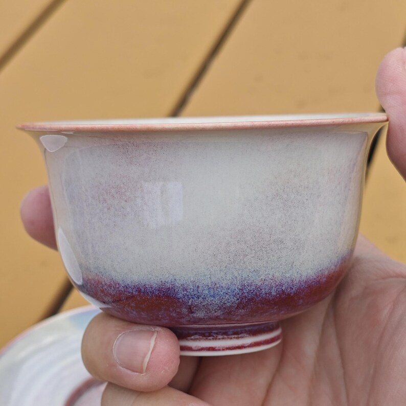 May include: A ceramic teacup with a white and purple glaze. The cup has a rounded shape with a flared rim and a small foot. The glaze transitions from white to purple near the base. The cup is held in a hand.