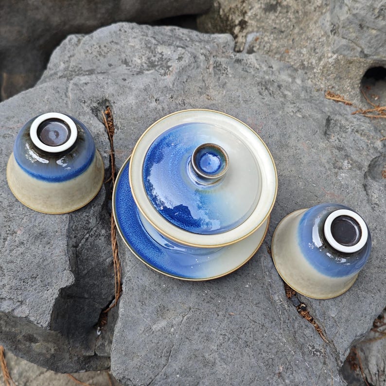 May include: A ceramic tea set consisting of a lidded gaiwan and two matching tea cups. Each piece features a gradient glaze transitioning from a deep cobalt blue at the top to a creamy off-white base, finished with a smooth, glossy texture.