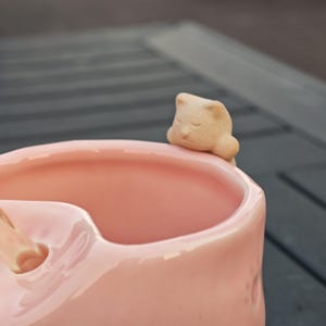 May include: A pink ceramic mug with a sleeping cat figurine attached to the rim. The cat is a light beige color, and the mug has a smooth, glossy finish. The mug is designed with a unique shape.