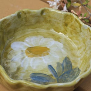May include: A ceramic bowl with a scalloped edge, painted in shades of yellow and green. The interior features a large white daisy with a yellow center and blue leaves. The bowl has a rustic, handmade appearance.