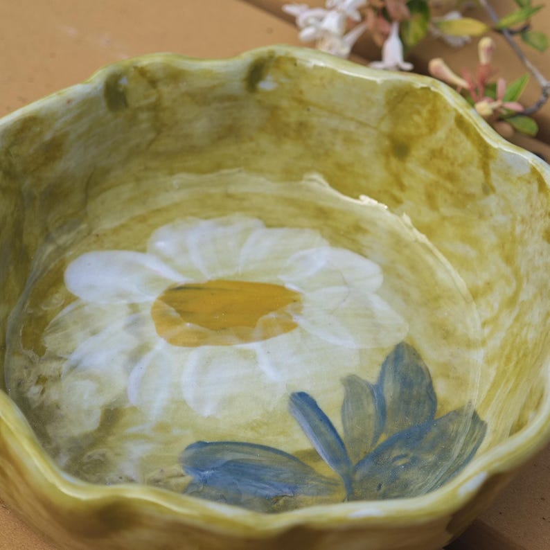 May include: A ceramic bowl with a scalloped edge, painted in shades of yellow and green. The interior features a large white daisy with a yellow center and blue leaves. The bowl has a rustic, handmade appearance.