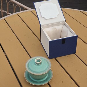 K&ouml;nnte beinhalten: Ein t&uuml;rkisfarbener Keramik-Gaiwan mit Deckel und Untertasse steht neben einer offenen, quadratischen Pr&auml;sentationsbox. Die Box hat eine tiefblaue Au&szlig;enseite, ein wei&szlig;es Innenfutter und einen wei&szlig;en rechteckigen Einsatz im Deckelinneren.