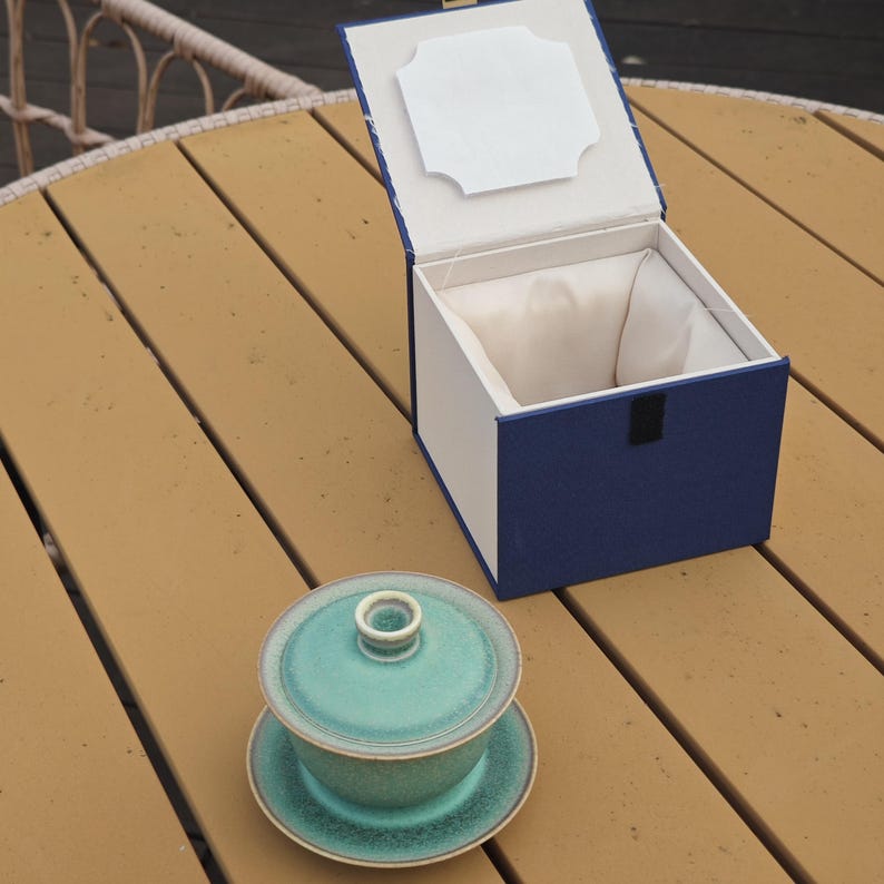 K&ouml;nnte beinhalten: Ein t&uuml;rkisfarbener Keramik-Gaiwan mit Deckel und Untertasse steht neben einer offenen, quadratischen Pr&auml;sentationsbox. Die Box hat eine tiefblaue Au&szlig;enseite, ein wei&szlig;es Innenfutter und einen wei&szlig;en rechteckigen Einsatz im Deckelinneren.
