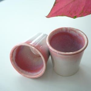 May include: Two ceramic cups with a gradient color scheme, transitioning from white to pink. The cups have a smooth, glossy finish and a slightly flared rim. The interior of the cups is a vibrant pink.