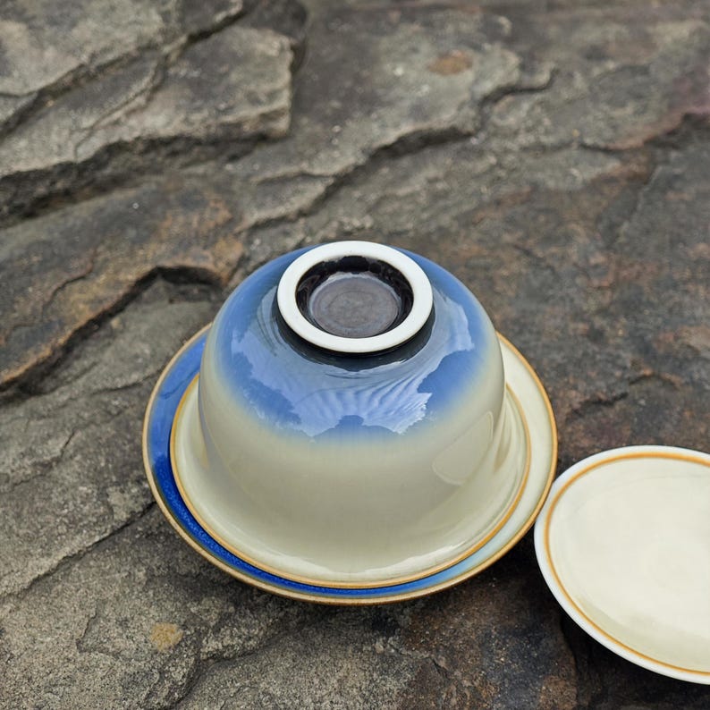 May include: A ceramic tea bowl and saucer set featuring a smooth, cream-colored base that transitions into a deep blue gradient toward the rim. The bowl is inverted on the saucer, revealing a dark circular foot ring with a white border.