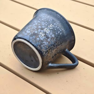 May include: A ceramic mug with a dark blue speckled glaze and a white interior rim. The mug has a curved handle and a Japanese inscription in white. The mug is resting on a wooden surface.