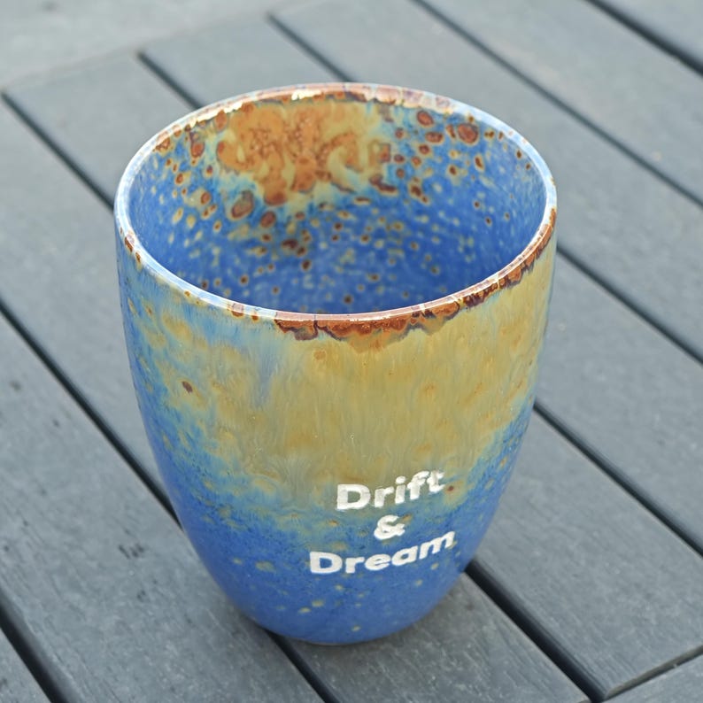 Puede incluir: Una taza de cer&aacute;mica con un esmalte azul y amarillo, con las palabras "Drift & Dream" en blanco. El borde tiene un acabado marr&oacute;n texturizado. La taza es cil&iacute;ndrica y est&aacute; sobre una superficie gris.