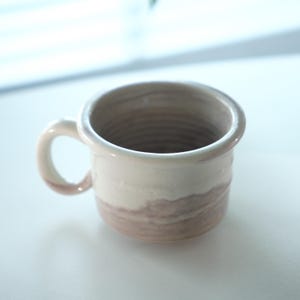 May include: A handmade ceramic mug with a rounded handle. The mug features a two-tone design, with a creamy white upper section and a muted brown lower section. The interior is a darker brown. The mug is suitable for coffee or tea.