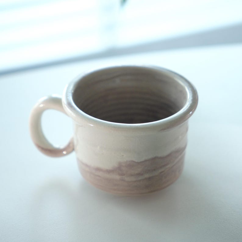 May include: A handmade ceramic mug with a rounded handle. The mug features a two-tone design, with a creamy white upper section and a muted brown lower section. The interior is a darker brown. The mug is suitable for coffee or tea.