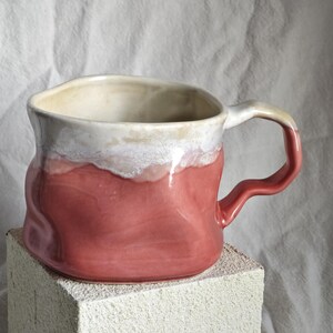May include: A handmade ceramic mug with a unique, organic shape. The mug features a gradient of colors, with a white rim, a pinkish-red body, and a cream-colored handle. The mug is sitting on a white block.