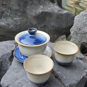 May include: A ceramic tea set consisting of a lidded gaiwan with a saucer and two matching tea cups. The pieces feature a cream-colored base with a deep blue gradient glaze on the lid, saucer, and the lower exterior sections of the cups.