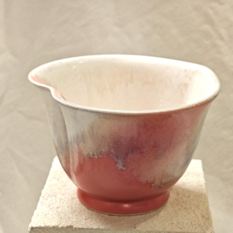 May include: A ceramic bowl with a flared rim and a footed base. The bowl features a gradient of colors, transitioning from a creamy white interior to a red base, with shades of pink and purple in between. The bowl is sitting on a light-colored surface.