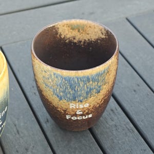 Puede incluir: Una taza de cer&aacute;mica con un esmalte marr&oacute;n, azul y beige. La taza tiene las palabras "Rise & Focus" en letras blancas en el lateral. La taza est&aacute; sobre una superficie de madera gris.