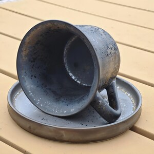 May include: A dark gray ceramic coffee cup and saucer set. The cup is overturned, revealing a speckled interior. The saucer is round with a slightly raised edge. The set has a rustic, handmade appearance.