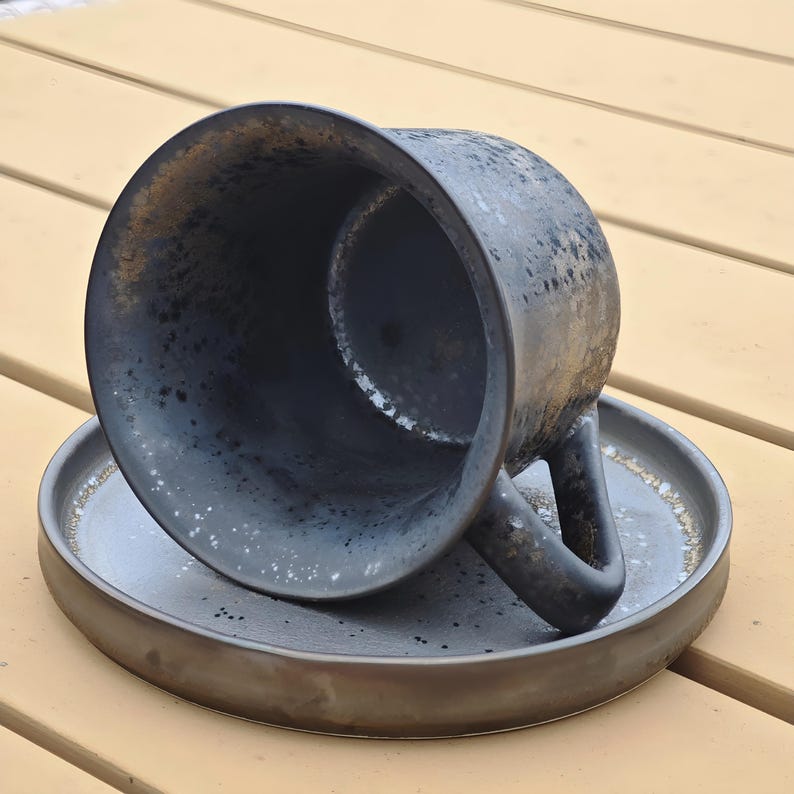 May include: A dark gray ceramic coffee cup and saucer set. The cup is overturned, revealing a speckled interior. The saucer is round with a slightly raised edge. The set has a rustic, handmade appearance.