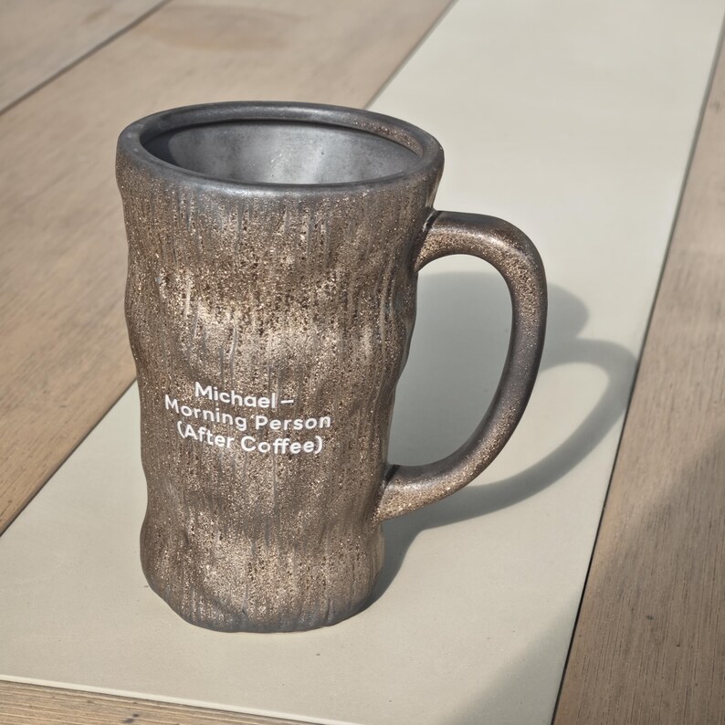 Puede incluir: Una taza de cer&aacute;mica cil&iacute;ndrica y alta con un exterior texturizado en tonos bronce met&aacute;lico y un interior liso de color gris oscuro. La taza cuenta con un asa y texto impreso en blanco que dice: Michael - Morning Person (After Coffee).