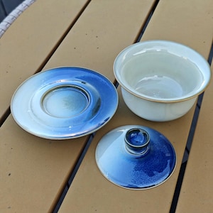 May include: A ceramic tea set consisting of a bowl, a saucer, and a lid. The bowl is off-white with a gold-toned rim. The saucer and lid feature a gradient design transitioning from a deep blue center to a lighter blue and white edge.