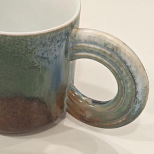 K&ouml;nnte beinhalten: Nahaufnahme einer Keramik-Tasse mit einem gro&szlig;en, gebogenen Henkel. Die Tasse hat einen Farbverlauf aus Gr&uuml;n, Blau und Braun. Der Innenraum ist wei&szlig;. Der Griff hat ein strukturiertes Design, und die Tasse scheint handgefertigt zu sein.