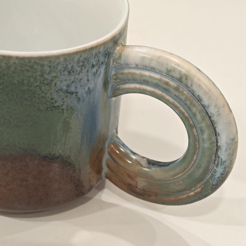 K&ouml;nnte beinhalten: Nahaufnahme einer Keramik-Tasse mit einem gro&szlig;en, gebogenen Henkel. Die Tasse hat einen Farbverlauf aus Gr&uuml;n, Blau und Braun. Der Innenraum ist wei&szlig;. Der Griff hat ein strukturiertes Design, und die Tasse scheint handgefertigt zu sein.